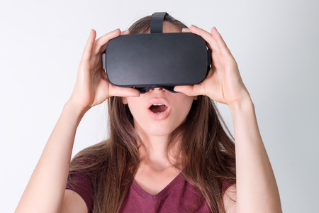 Amazed positive woman wearing virtual reality goggles headset, vr box. Connection, technology, new generation, progress concept. Girl surprised by something in virtual reality. Studio shot on grayの写真素材