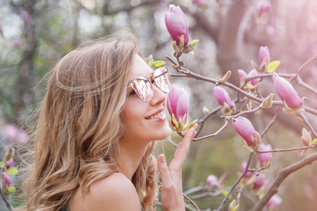 Young blonde woman near blossoming magnolia flowers tree in spring park on sunny day. Magnolia trees. Beautiful happy girl enjoying smell in a flowering spring gardenの写真素材