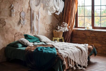 Stylish room interior with large comfortable bed. Beige and white dream catchers and feathers hanging above gypsy or hippie slyle bed in dark autumn bedroom interiorの写真素材