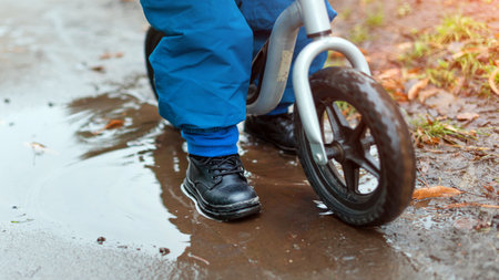 A child is riding a balance bike through a wet and muddy path with puddles. The image captures a playful and carefree childhood moment enjoying outdoor activities despite the conditionsの写真素材