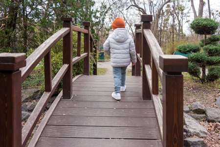 A young child dressed in warm clothing crosses a wooden bridge surrounded by lush greenery. The photograph captures a peaceful and natural outdoor setting perfect for family and exploration themesの写真素材