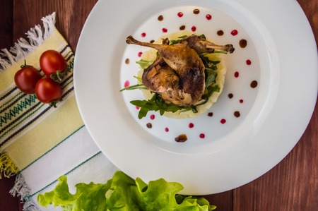 Roasted quail on a bed with mashed potatoes, rocket leaves and berry sauce. On a white plate and wooden rustic background. Top viewの写真素材