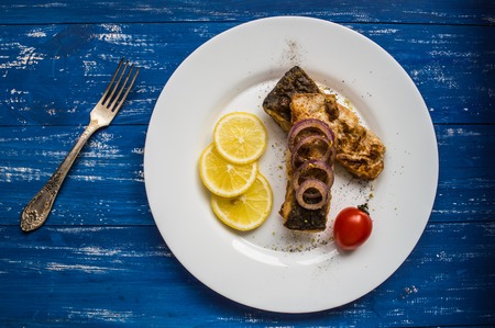 Fried fish fillet with lemon and tomatoes on a white plate. Wooden blue background