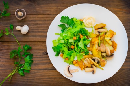 Fried potatoes roasted with wild mushrooms on white plate and green lettuce on wooden table...
