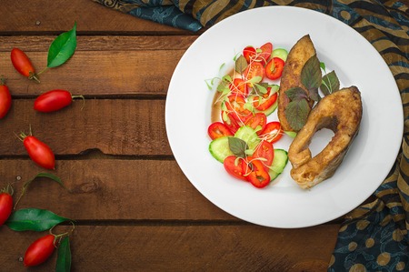 Grilled fish steak with vegetables on plate: tomatoes, microgran, cucumber, tasty and healthy dinner. Wooden rustic background. Top view.の写真素材
