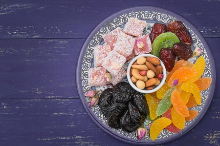 Set of eastern desserts. Marmalade, kivi, prunes, rahat lokum, nuts, mandarin, persimmon, dried apricots, pistachios, dates, raisins in a colorful blue plate Wooden rustic background Top viewの写真素材