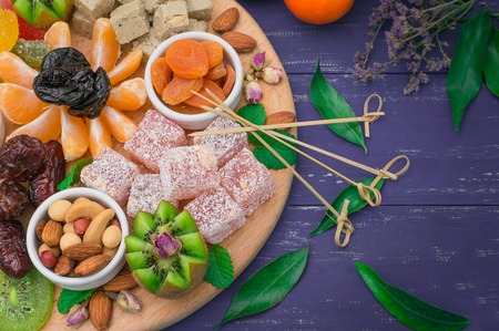 Set of eastern desserts. Marmalade, kivi, prunes, rahat lokum, nuts, mandarin, persimmon, dried apricots, pistachios, dates raisins in a colorful blue Wooden rustic background Top viewの写真素材