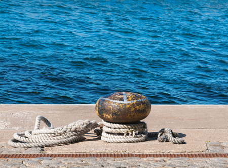 Rusty yellow bollard with worn out rope against blue sea; Empty mooring at the portの写真素材