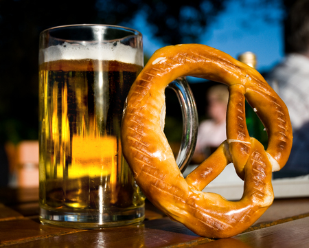 A glass of beer and a pretzel on a table in closeup; Hearty snack; Visit at beer garden in the evening; Alcoholic beverage and special breadの写真素材