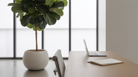A modern office setup featuring a laptop and notebook on a wooden desk, with a large fiddle leaf fig plant by the window.の素材