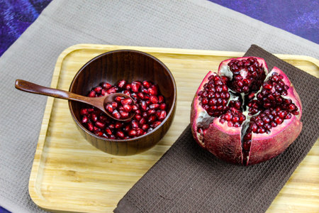 Wooden bowl containing wooden spoon and red pomegranate seeds. Pomegranate fruit open divided into five parts held together on a napkin / cloth on wooden tray on a blue background. Fruit pomegranate.の写真素材