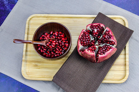 Wooden bowl containing wooden spoon and red pomegranate seeds. Pomegranate fruit open divided into five parts held together on a napkin / cloth on wooden tray on a blue background. Fruit pomegranate.の写真素材