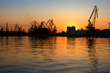 Beautiful sunset behind Seaport Varna, Bulgaria. Silhouettes of industrial cranes and buildings with reflections in the waterの写真素材
