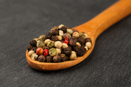 Macro shot of black, green, white and red peppercorn mix in a wooden spoon on a dark stone kitchen board. Shallow depth of fieldの写真素材