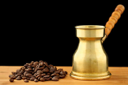 Old, vintage brass turkish coffee pot (dzhezve) with carved wooden handle and a pile of roasted coffee beans on a wooden table against black background. Space for textの写真素材