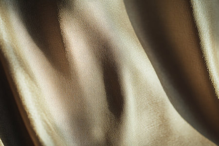 A textured golden fabric undulates, displaying an abstract pattern accentuated by natural light and soft shadows. Subtle variations in tone add depth and visual interest.の素材