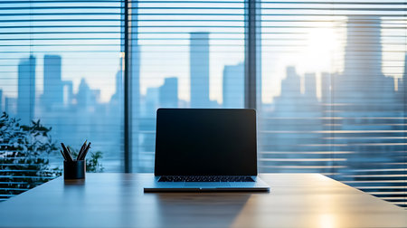 Silver laptop centered on a wooden desk, pens in a container, with a high-rise city skyline view behind blue blinds. Sunshine filters through the window.の素材