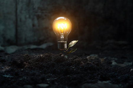 Light bulb emitting warm glow from the soil on top of which a green plant is seen growing against a textured concrete wall background.の素材