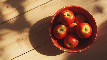 Top-down view showing a red bowl full of ripe red apples set on a light wooden surface. Sunlight creates strong shadows, enhancing the fresh, rustic feel.の素材