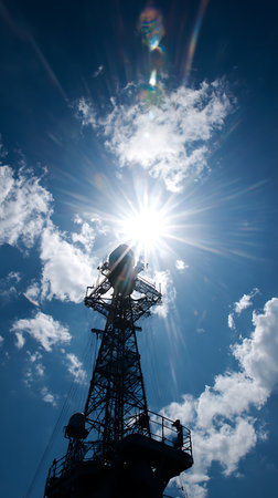 Silhouette of a ship's communications mast against a vibrant blue sky with scattered clouds. The sun shines brightly behind the tower, creating a starburst effect and lens flare.の素材