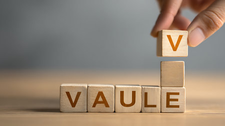 Close up depicts a hand placing a wooden block with the letter V on top of wooden blocks that spell VAULE on a wooden surface against a grey background.の素材