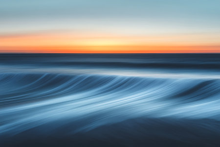 Dynamic long-exposure photography reveals smooth ocean waves under a vibrant orange and blue sunset. The blurred motion creates an abstract, serene waterscape.の素材