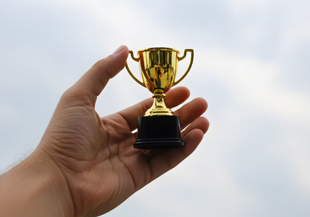 Hand holding a gold trophy cup on blue sky background, success conceptの素材