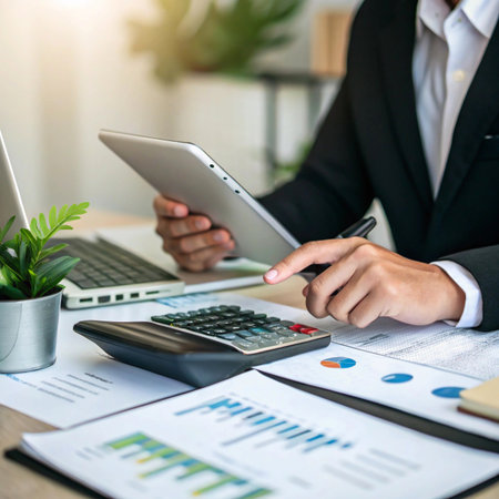 A person in a suit uses a tablet and calculator to review financial charts and documents at a desk with a laptop.の素材