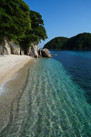 A serene tropical beach with incredibly clear turquoise water, white sand, and lush green hills under a bright blue sky.の素材
