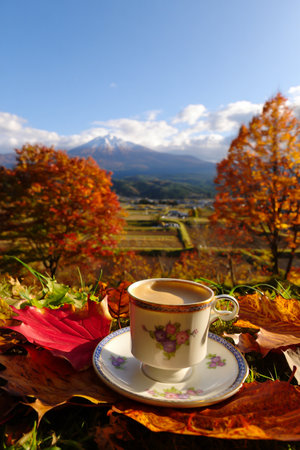 A warm cup of coffee sits on a table adorned with autumn leaves, overlooking a scenic mountain landscape.の素材
