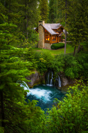 A warm, inviting cabin glows with light beside a vibrant blue waterfall surrounded by dense, verdant forest.の素材
