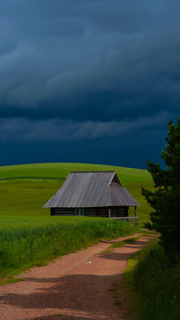 A lone barn sits in a vast green field under a dark, ominous, and cloudy sky, suggesting an approaching storm.の素材