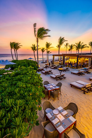 A beautiful beachside restaurant with tables set on sand, overlooking the ocean at sunset with palm trees.の素材