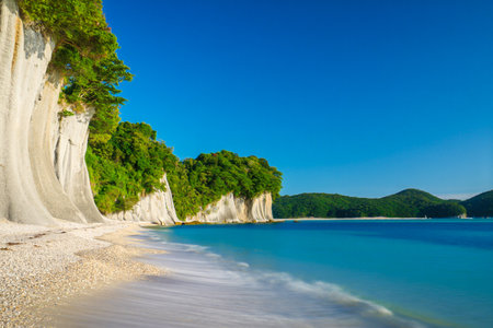 A serene tropical beach with dramatic white cliffs, lush green foliage, and crystal clear turquoise water under a bright blue sky.の素材