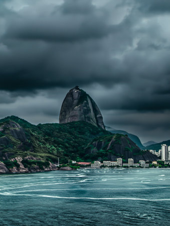 Dramatic dark clouds loom over the iconic Sugarloaf Mountain and the waters of Guanabara Bay, with city buildings visible.の素材