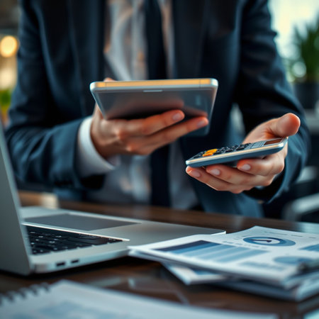 Businessman using smartphone and calculator at desk with laptop and documentsの素材
