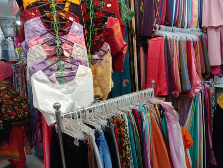 This image captures a vibrant market stall showcasing a variety of colorful garments. The stall features an array of tops and dresses in bright hues such as pink, purple, blue, and orange. The garments are neatly arranged on hangers and racks, creating an eye-catching display. The foreground prominently displays a white top with intricate designs, while the background reveals more colorful clothing items. The overall atmosphere is lively and bustling, typical of a busy market setting.の写真素材