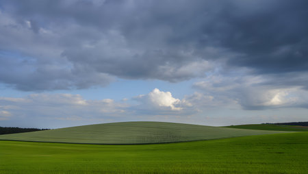 A wide-angle view captures a vibrant green rolling hill under a dramatic sky filled with dark, textured storm clouds. Patches of lighter sky peek through, creating a sense of impending weather.の素材