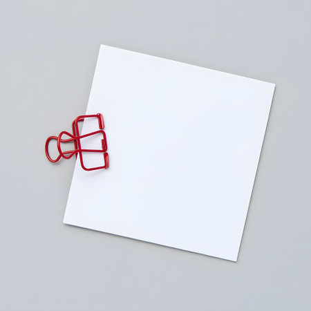 Blank white square note paper held by a red binder clip on a gray surfaceの素材