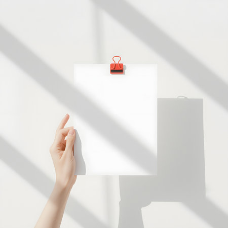 Hand holding blank paper with red clip against white wall with diagonal shadow patternsの素材