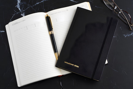 A sophisticated flat lay featuring a closed black notebook and an open white journal, a pen resting between the pages, on a dark marbled background.の素材