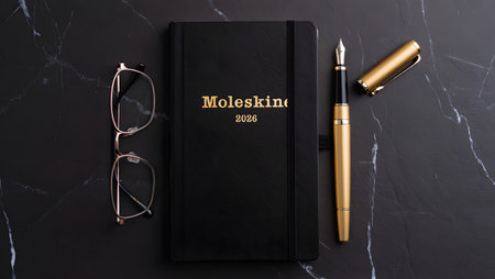 A flat lay composition featuring a black Moleskine notebook, eyeglasses, and a wooden fountain pen on a dark, textured background.の素材
