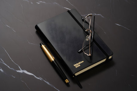 A sophisticated flat lay featuring a closed black notebook, a gold pen, and reading glasses resting on a dark marble background.の素材
