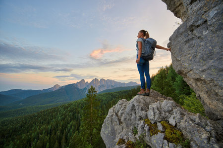 A lone hiker stands on a rocky cliff overlooking a vast forest and mountain range.の素材