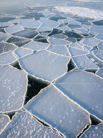 This image captures the intricate pattern of cracks on the surface of a frozen lake. The ice is segmented into various geometric shapes, with some sections covered in a light dusting of snow. The cracks create a striking contrast against the smooth, icy surface, highlighting the natural beauty of winter landscapes.の素材