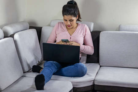 Young woman with smartphone using laptop at homeの写真素材
