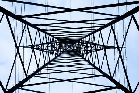 View from the bottom upwards to steel tower of electric main or electricity transmission line with the wires silhouette on background of cloudy blue skyの写真素材