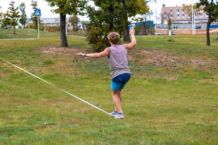 A young man in a public park learning to walk a tightrope. Slacklining is a practice in balance that typically uses nylon or polyester webbing tensioned between two anchor points.の写真素材