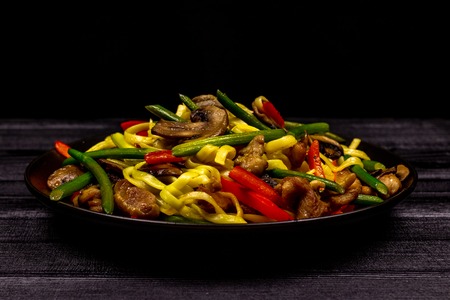 Appetizing stir fried noodles with garlic sprouts, mushrooms, red pepper and chicken meat on a black rustic background of a wooden table. A popular Asian dishの写真素材