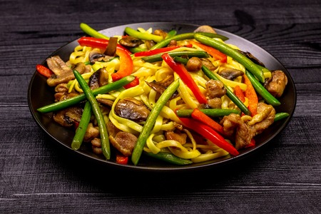 Appetizing stir fried noodles with garlic sprouts, mushrooms, red pepper and chicken meat on a black rustic background of a wooden table. A popular Asian dish.の写真素材